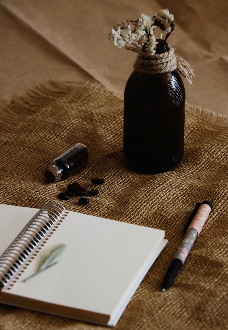 An open notebook and pen beside dried flowers in a dark glass bottle