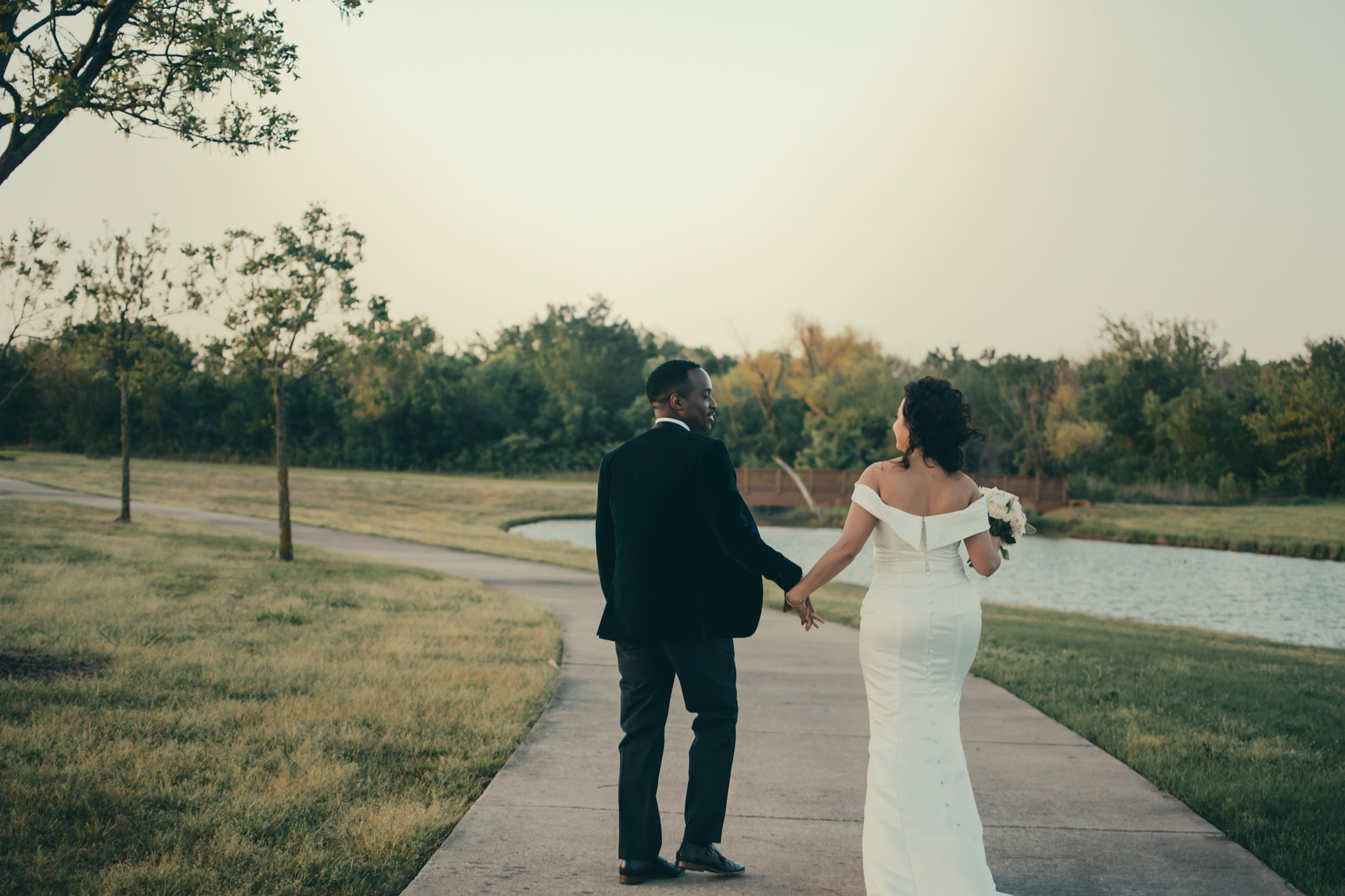 A newly married couple holding hands on a path beside a pond at dusk